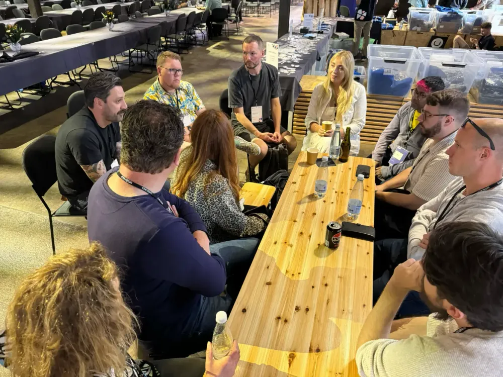 A wooden picnic bench style table surrounded by people. Some are facing the camera and some have their backs to the camera.